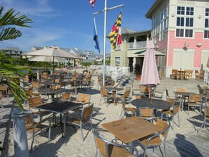 A beachfront patio with tables and chairs, a flagpole with flags, and a building with a balcony in the background.