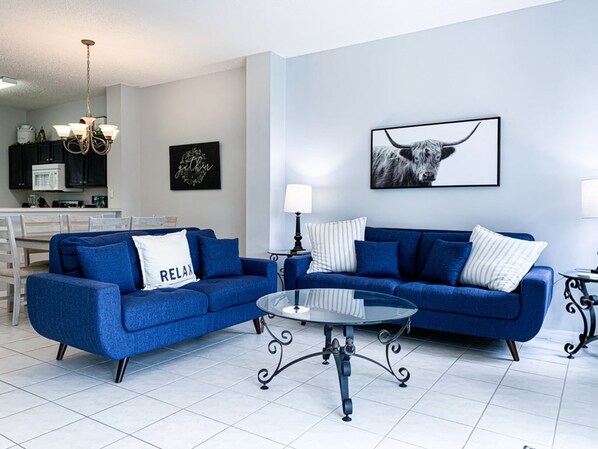 A living room with a blue sofa set, a glass coffee table, and a kitchen in the background. - Luxury Lake View 3 bedroom in resort community. Disney 3 miles (Kissimmee)