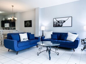 Un salon avec un canapé bleu, une table basse en verre et une cuisine en arrière-plan.