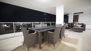 A modern outdoor dining area with a glass-top table and wicker chairs, overlooking a cityscape at night. - PENTHOUSE STYLE 4 BEDROOMS! (Bundall)