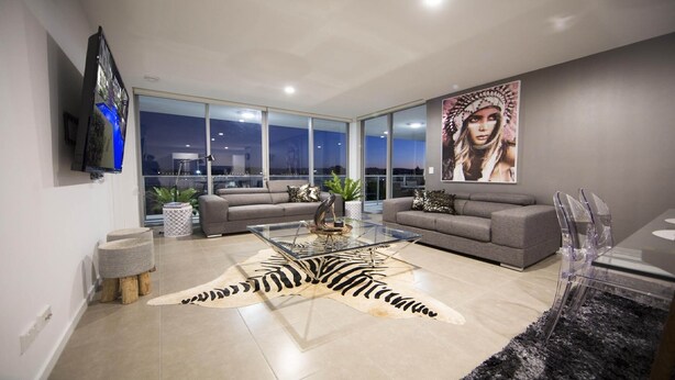 A modern living room with a glass coffee table, two grey sofas, a zebra print rug, and a large window offering a city view. - PENTHOUSE STYLE 4 BEDROOMS! (Bundall)