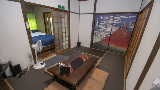 A traditional Japanese-style room with tatami flooring, a low table with cushions, a bed with a blue blanket, and a wall with a mountain painting. - Ra Villa Hanazono (Osaka)