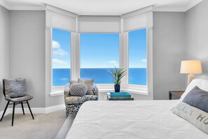 A bedroom with a large bay window, a bed, a chair, a nightstand, and a lamp.