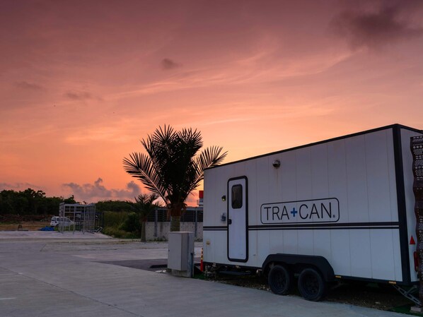 A white trailer with "TRA+CAN" branding parked in a lot with a palm tree and a building in the background. - T-PARK Airport Side 1 (Miyakojima)