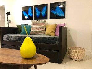 A modern living room with a sofa, colorful cushions, a yellow vase on a wooden table, and three butterfly paintings on the wall.