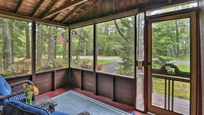 A covered porch with large windows, a wicker chair, a small table with flowers, and a view of a forested area. - 'Sprucewold' Boothbay Harbor Cottage w/ Deck (Boothbay Harbor)