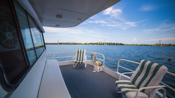 A boat deck with two striped lounge chairs, a steering wheel, and a view of a calm water body with distant land. - Happy’s Houseboat Key West (Key West)