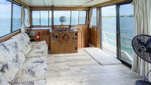 A cabin with a wooden control panel, a sofa, and large windows offering a view of the water. - Happy’s Houseboat Key West (Key West)