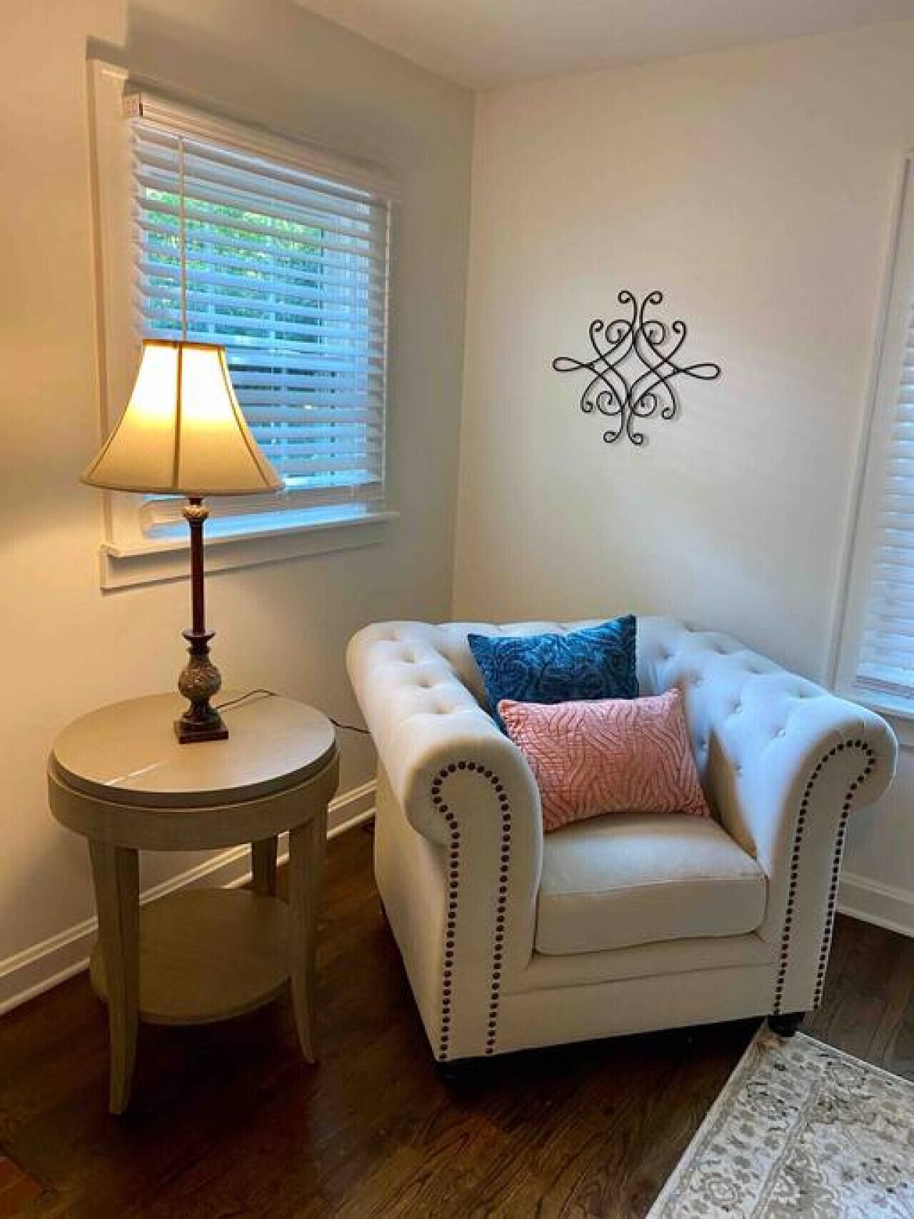 A room with a white armchair, a round side table with a lamp, and a decorative wall piece.