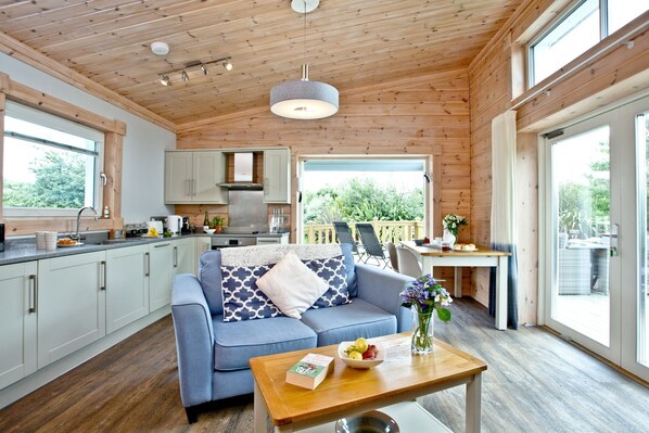 A modern kitchen with grey cabinets, a blue sofa, and a wooden table with a book and fruit. - Swanpool, Great Field Lodges -  a lodge that sleeps 2 guests  in 1 bedroom (Braunton)