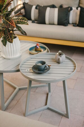 Uma mesa redonda com um bule, xícara, pires e um pequeno prato com sobremesa. Um vaso de planta está ao fundo, e um sofá com almofadas é visível.