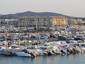 A marina with numerous boats docked, a parking area with cars, and a building complex in the background.