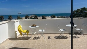 Un balcón con vistas a la playa, sillas plegables blancas, una silla amarilla y una mesa con un sombrero.