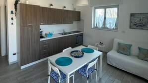 A modern kitchen with wooden cabinets, a dining table with chairs, a white sofa, and a window with curtains. - SoleeMare Barletta Apartments: Ocean View Suite (Barletta)