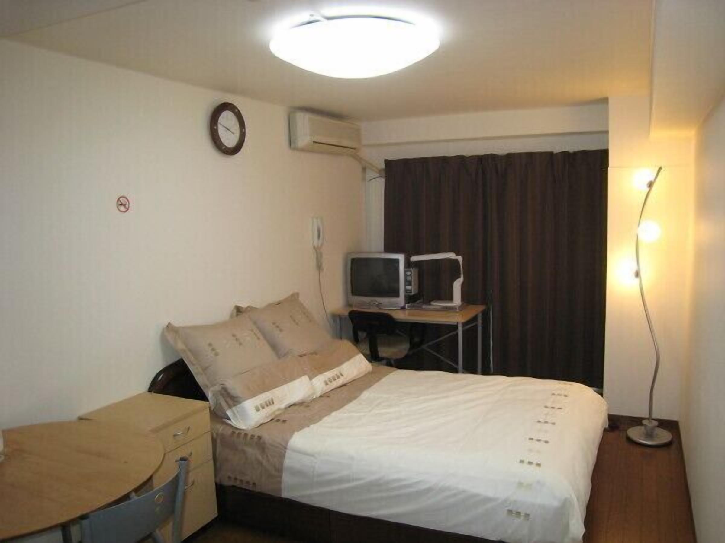 A hotel room with a bed, a desk, a television, an air conditioning unit, a clock, and a lamp.