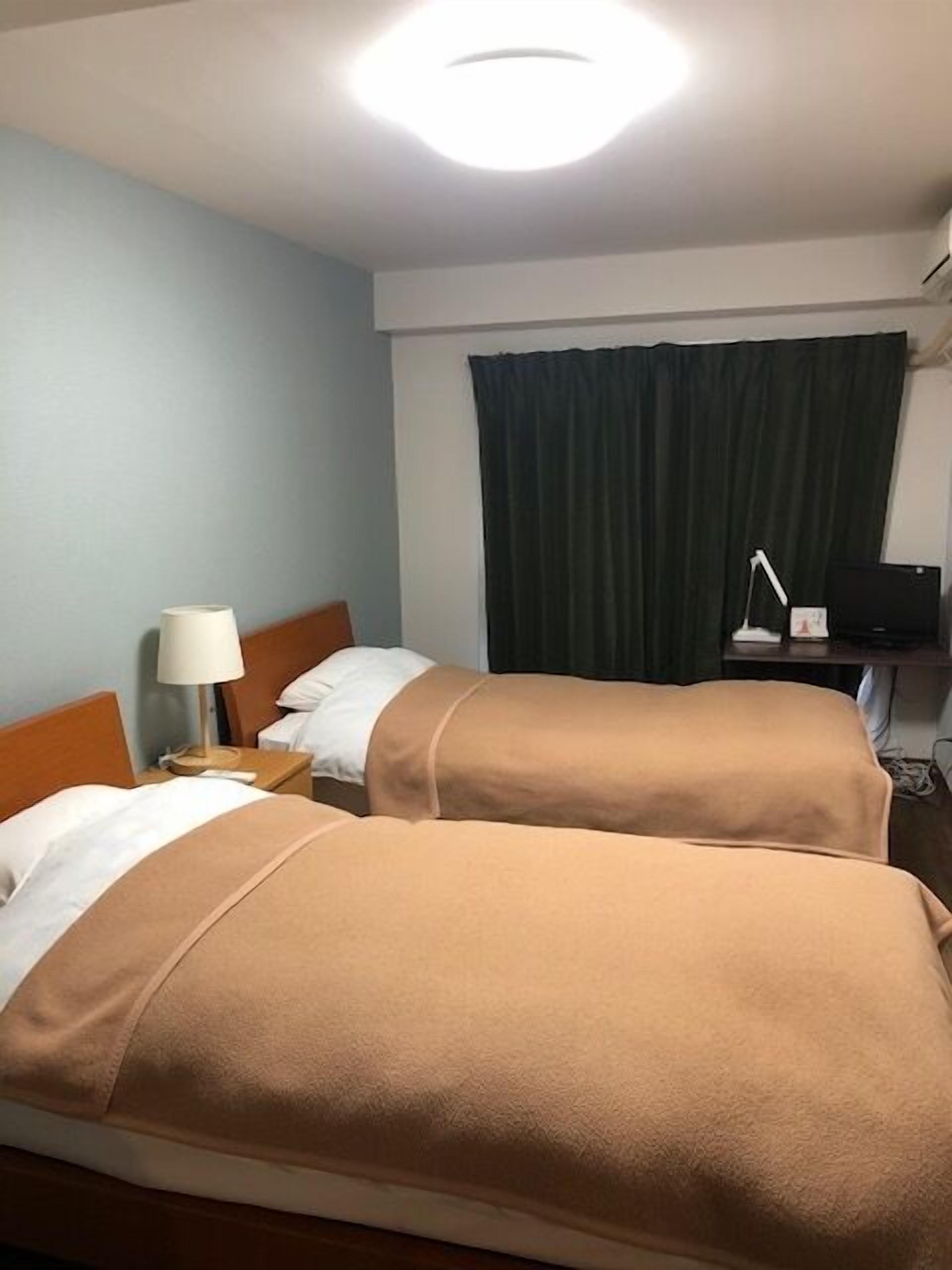 A hotel room with two beds, a desk, a lamp, a TV, and a curtain.