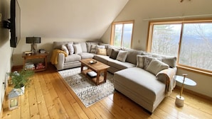 Un salon chaleureux, aménagé dans un style industriel, avec un grand canapé d’angle, une table basse en bois et une petite table d’appoint. Une grande fenêtre offre une vue sur l’extérieur.
