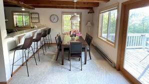 Un espace repas doté d’un plafond en bois, d’une table à manger avec des chaises, d’un vase rempli de fleurs et de grandes fenêtres.