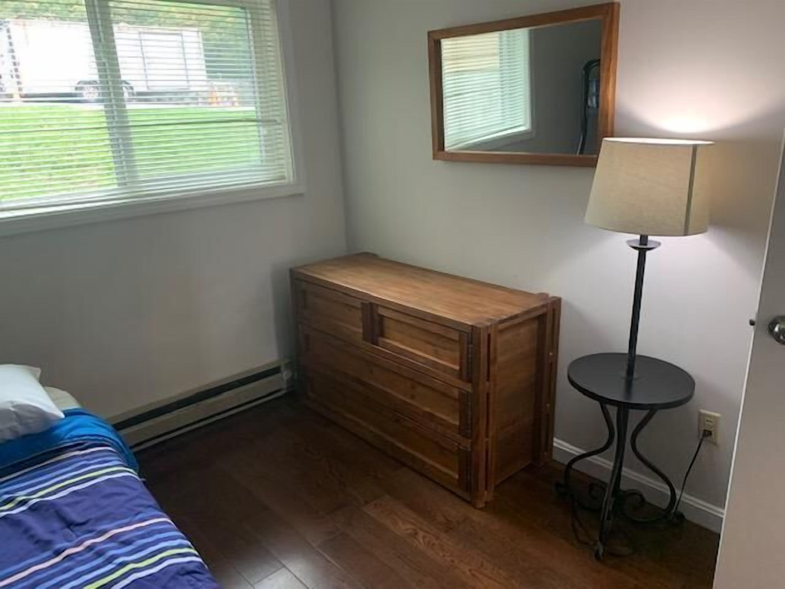 A bedroom with a wooden dresser, a bed with a striped bedspread, a standing lamp, and a window with blinds.