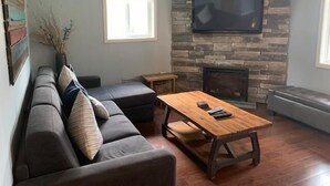 Ein Wohnzimmer mit Kamin aus Stein, einem darüber montierten Flachbildfernseher, einem dunkelgrauen Sofa mit Kissen und einem Couchtisch aus Holz.