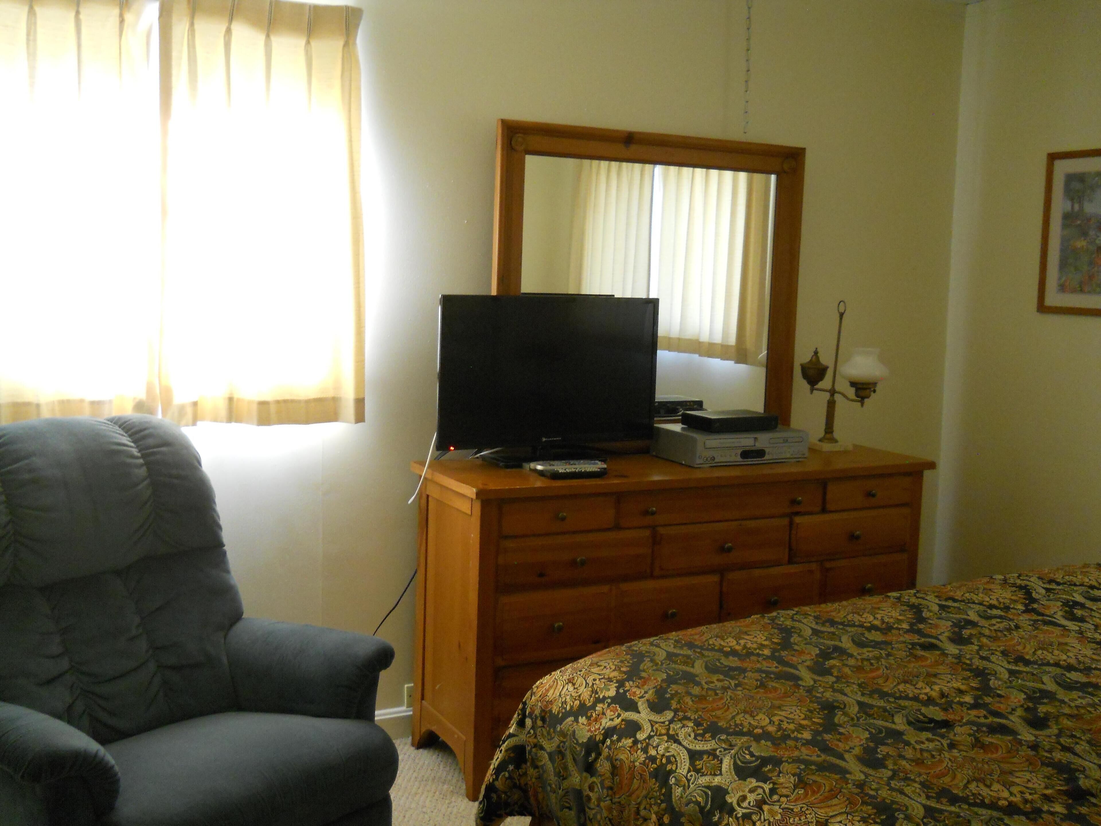 Un dormitorio con cama, cómoda con televisor, una silla y ventana con cortinas.