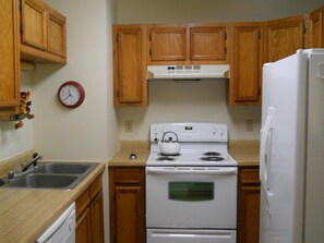 Una cocina con gabinetes de madera, una estufa blanca, un refrigerador, un lavamanos y un reloj en la pared.