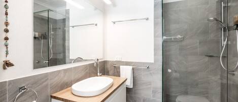 Une salle de bain moderne avec une douche à parois de verre, un lavabo blanc doté d’un plan de toilette en bois et un grand miroir.