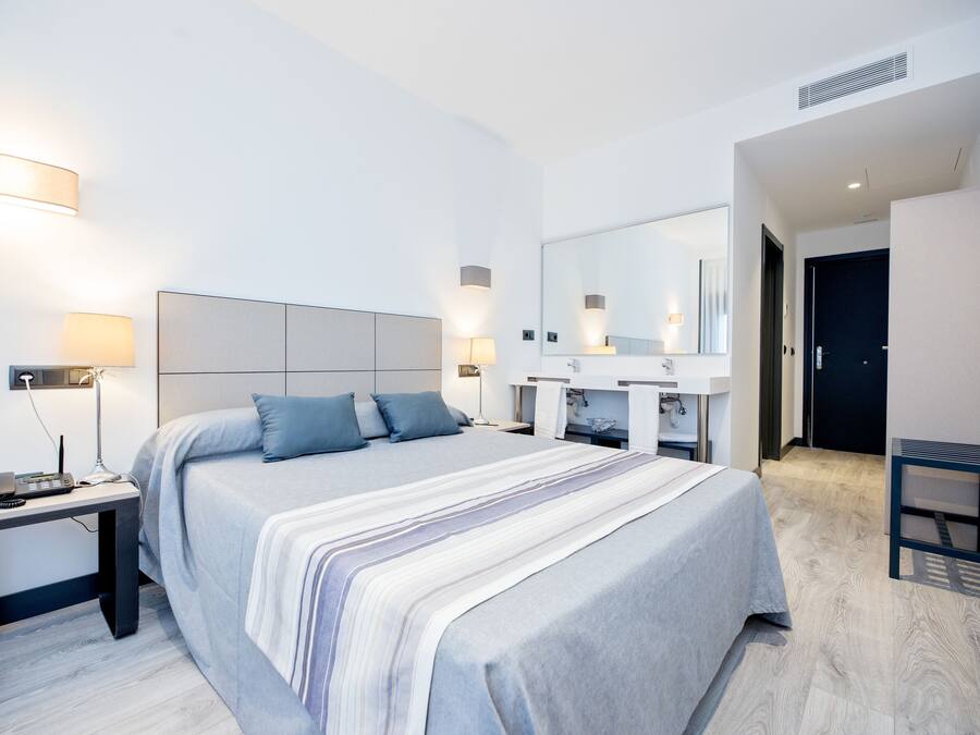 Una habitación de hotel moderna con una cama grande, mesitas de noche, un espejo y un baño con lavamanos y espejo.