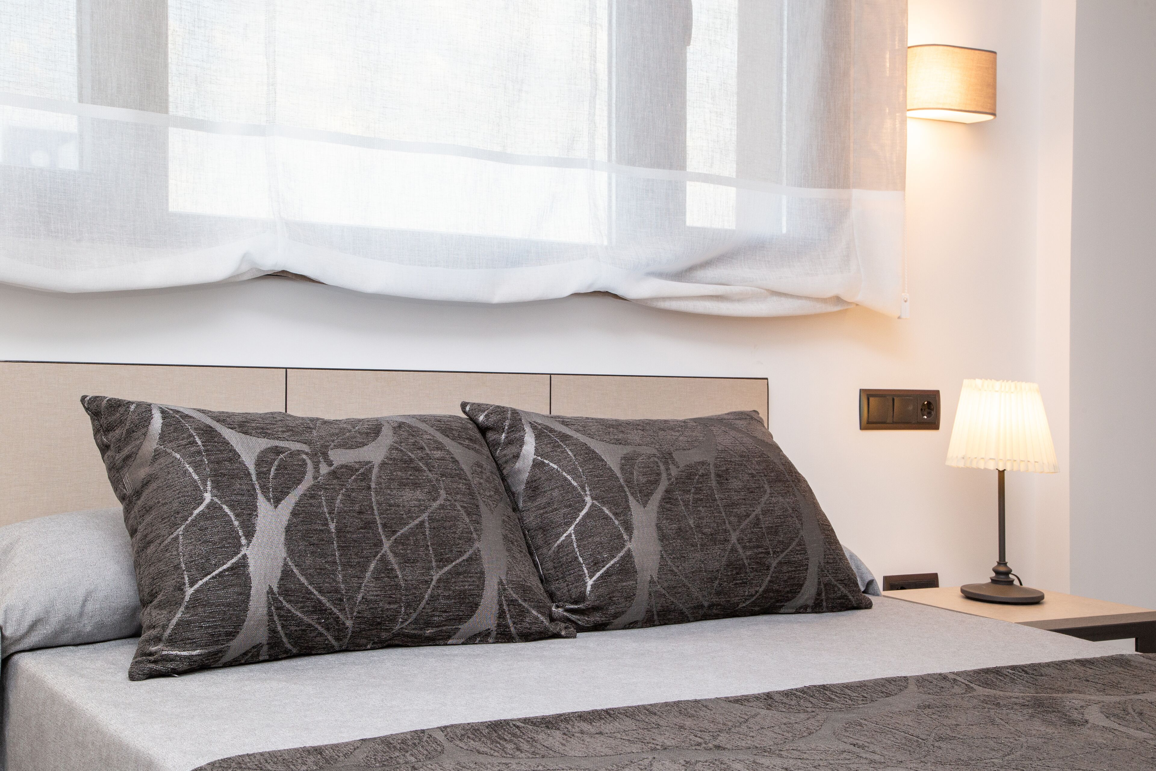 A neatly made bed with two gray pillows, a bedside lamp, and a window with sheer curtains.