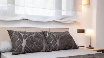 A neatly made bed with two gray pillows, a bedside lamp, and a window with sheer curtains.