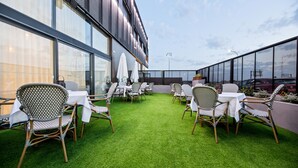Un espacio al aire libre con sillas y mesas de mimbre, césped artificial verde y un edificio moderno con grandes ventanales.