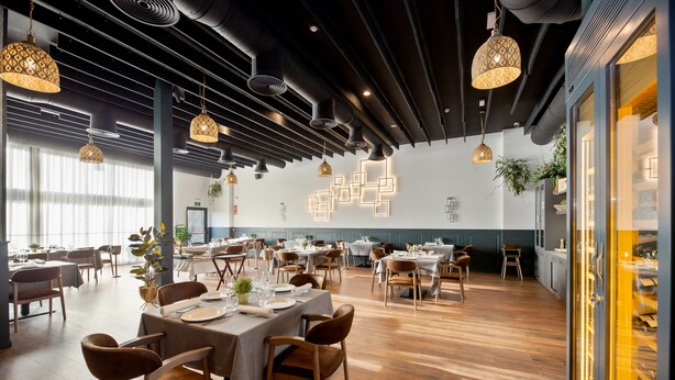 Un restaurante moderno con suelo de madera, mesas dispuestas con manteles blancos y lámparas colgantes.
