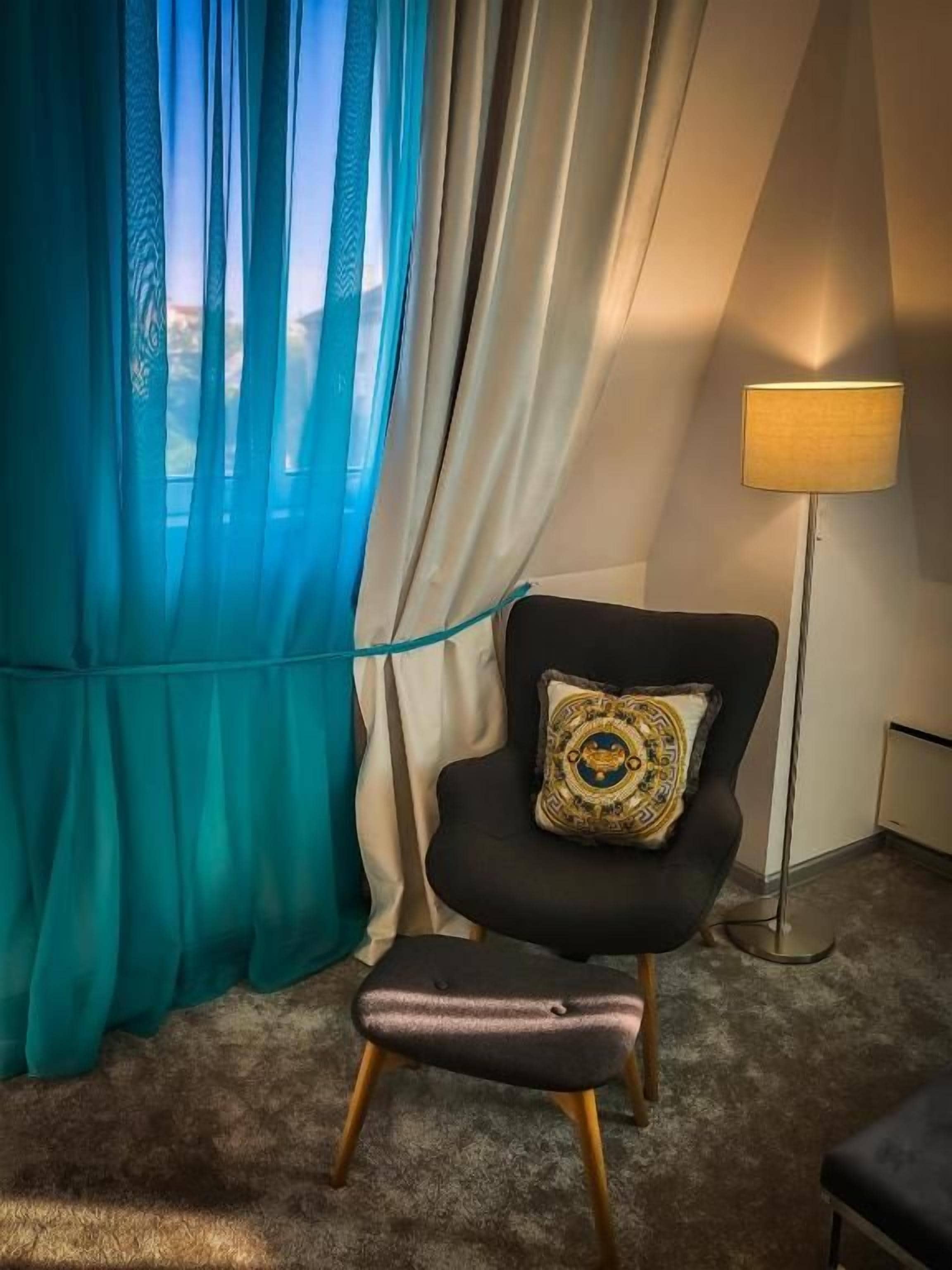 Una habitación con un sillón negro, un pequeño otoman, una lámpara de pie y vistas a través de cortinas translúcidas.