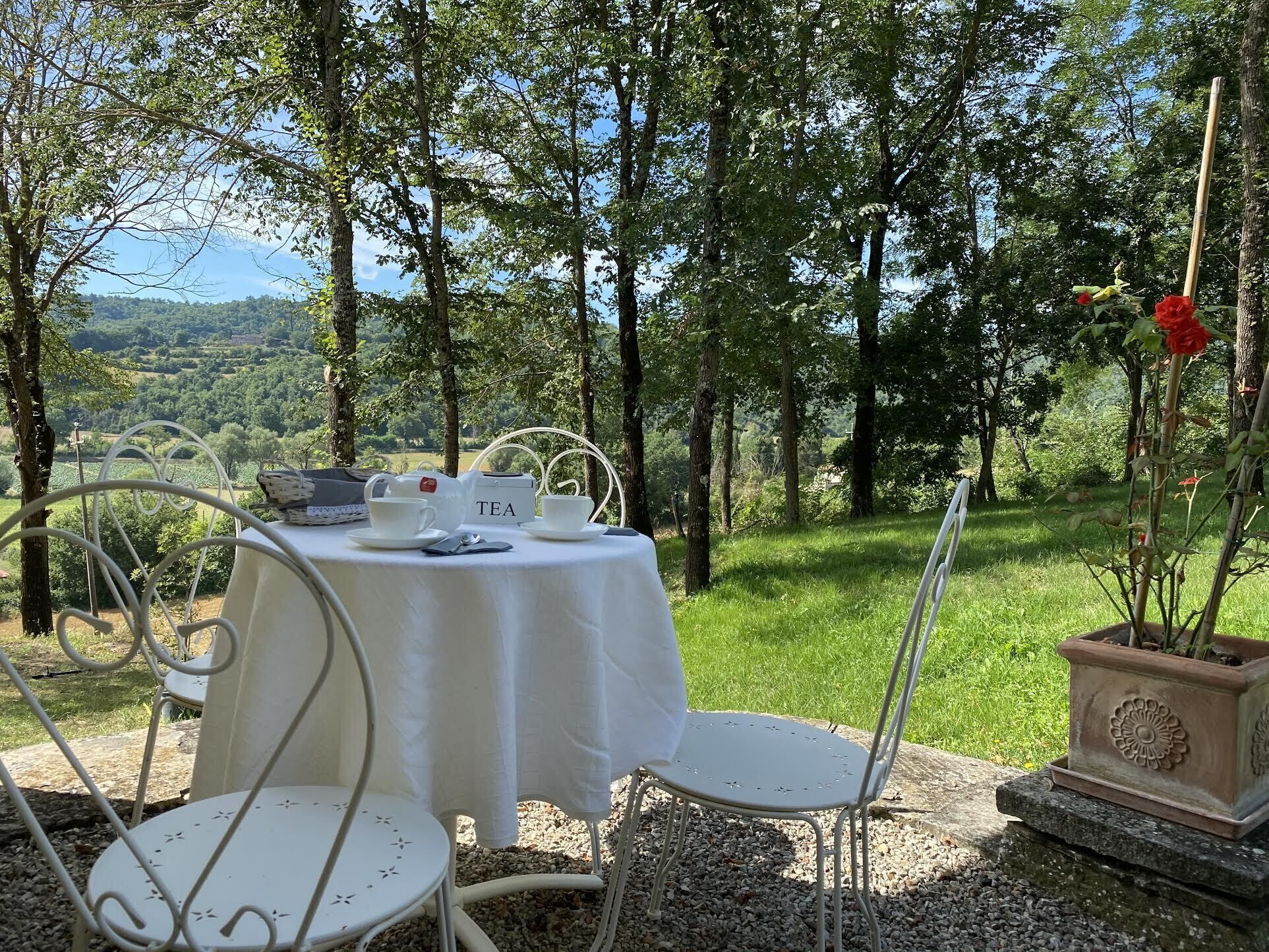 An outdoor dining area with a table set for tea, surrounded by white chairs and a view of a lush landscape.