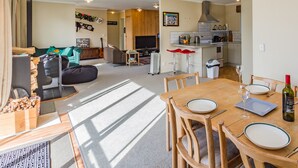 Un vaste espace de vie comprenant un coin repas avec une table pour deux, une cuisine à l’arrière-plan et une grande fenêtre laissant entrer la lumière naturelle.