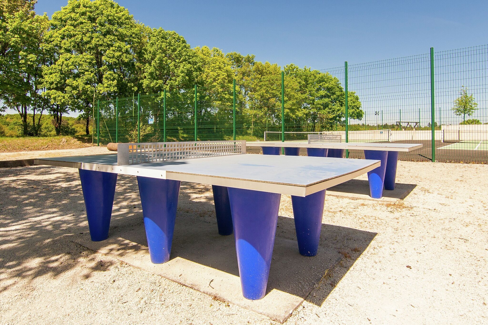 Een tafeltennistafel met blauwe poten, omringd door een groene hek, in een buitenruimte met bomen en een heldere lucht.