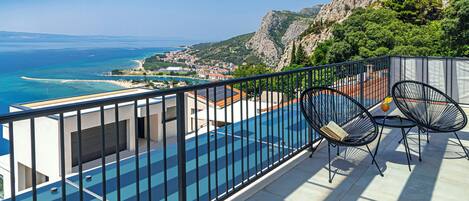 A balcony with a pool and chairs overlooking a coastal town and mountains.