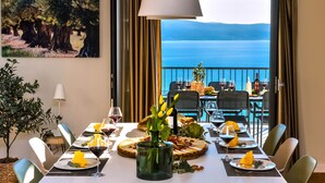 Een eettafel gedekt voor zes personen, met uitzicht op zee, een schilderij aan de muur en een hangende lamp.