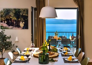 A dining table set for six with a view of the sea, a painting on the wall, and a hanging light fixture. - House Villa The View (Omis)
