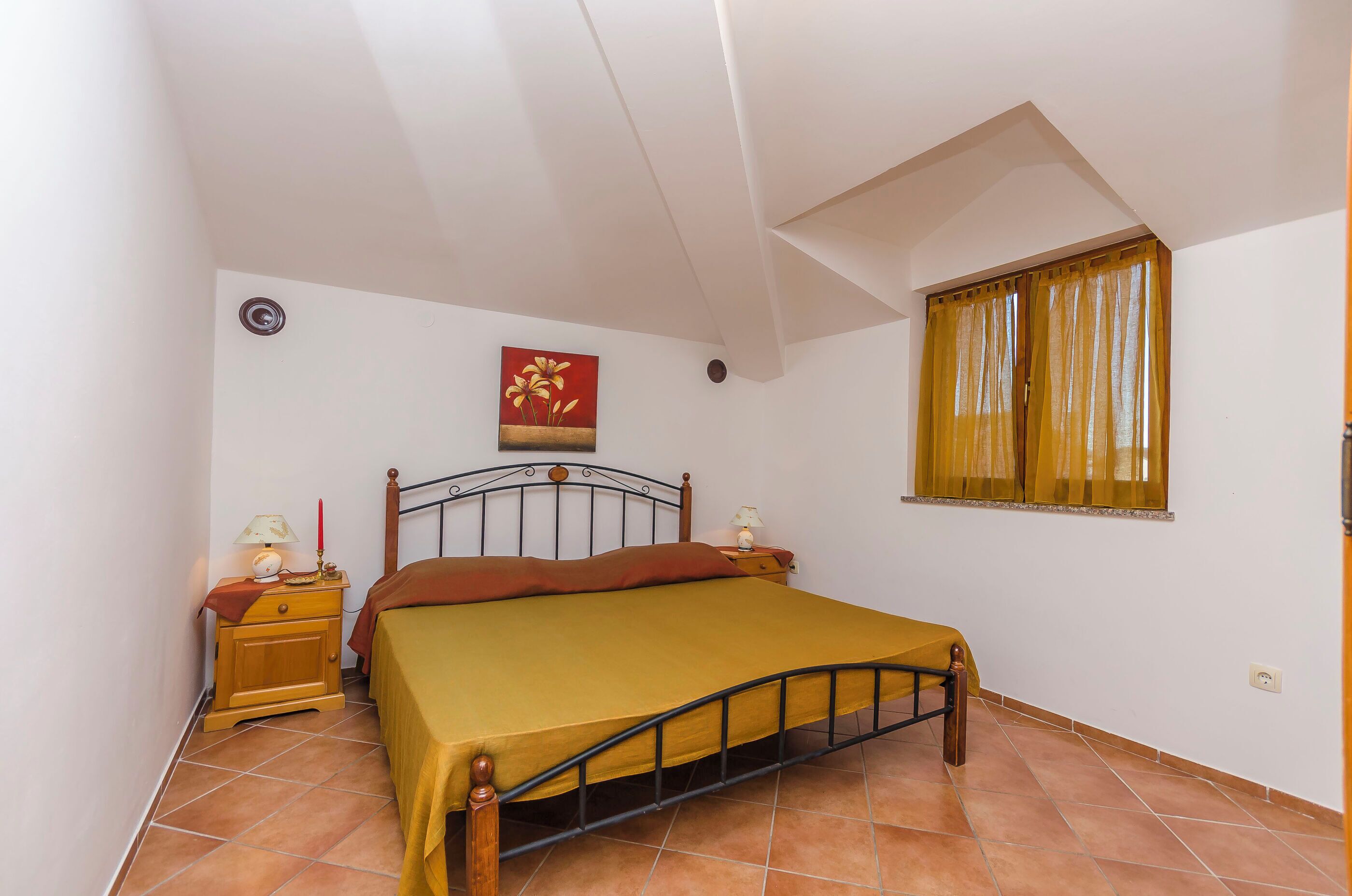 Quarto com cama, mesa de cabeceira, abajur, quadro e janela com cortinas.