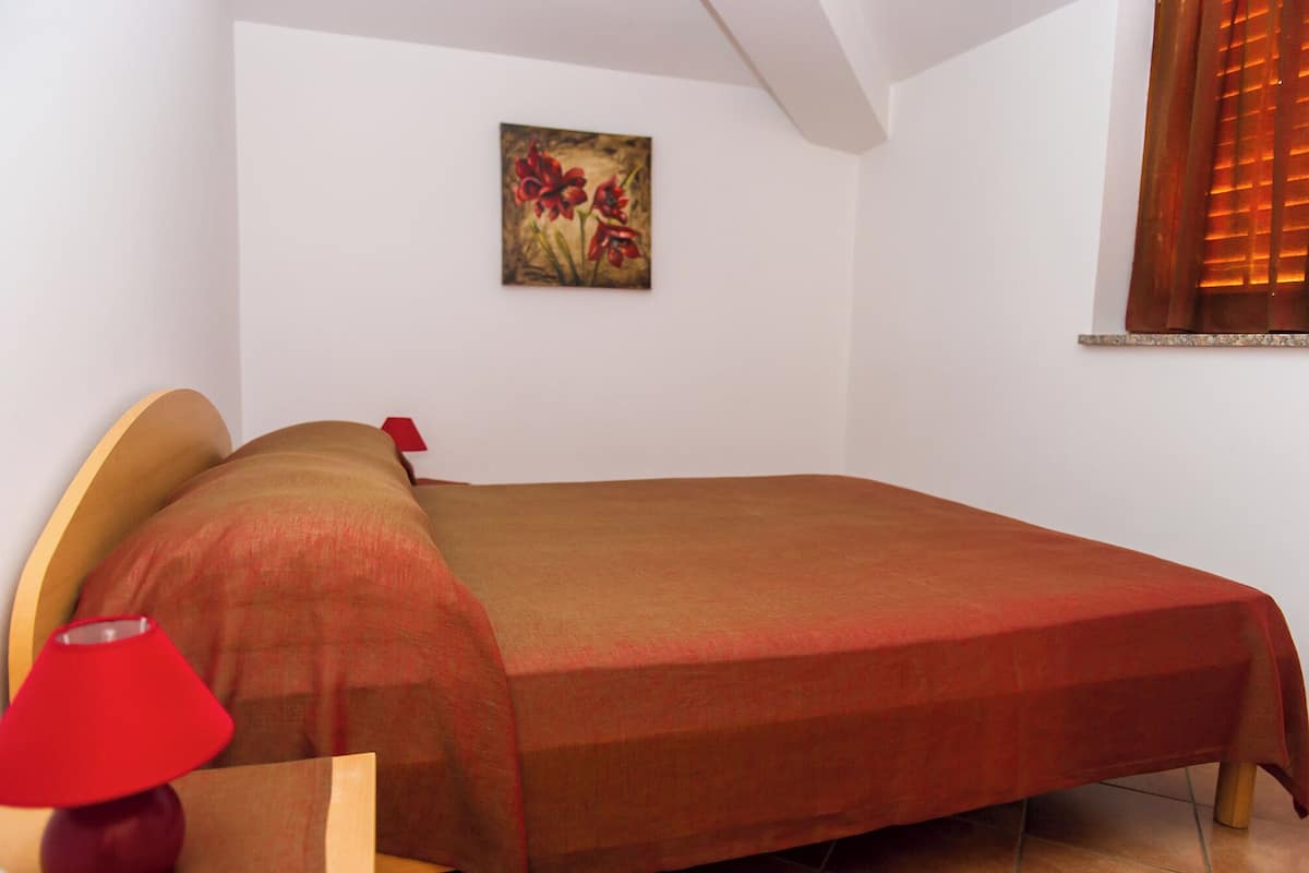 A neatly made bed with a red blanket and a red lamp beside it.