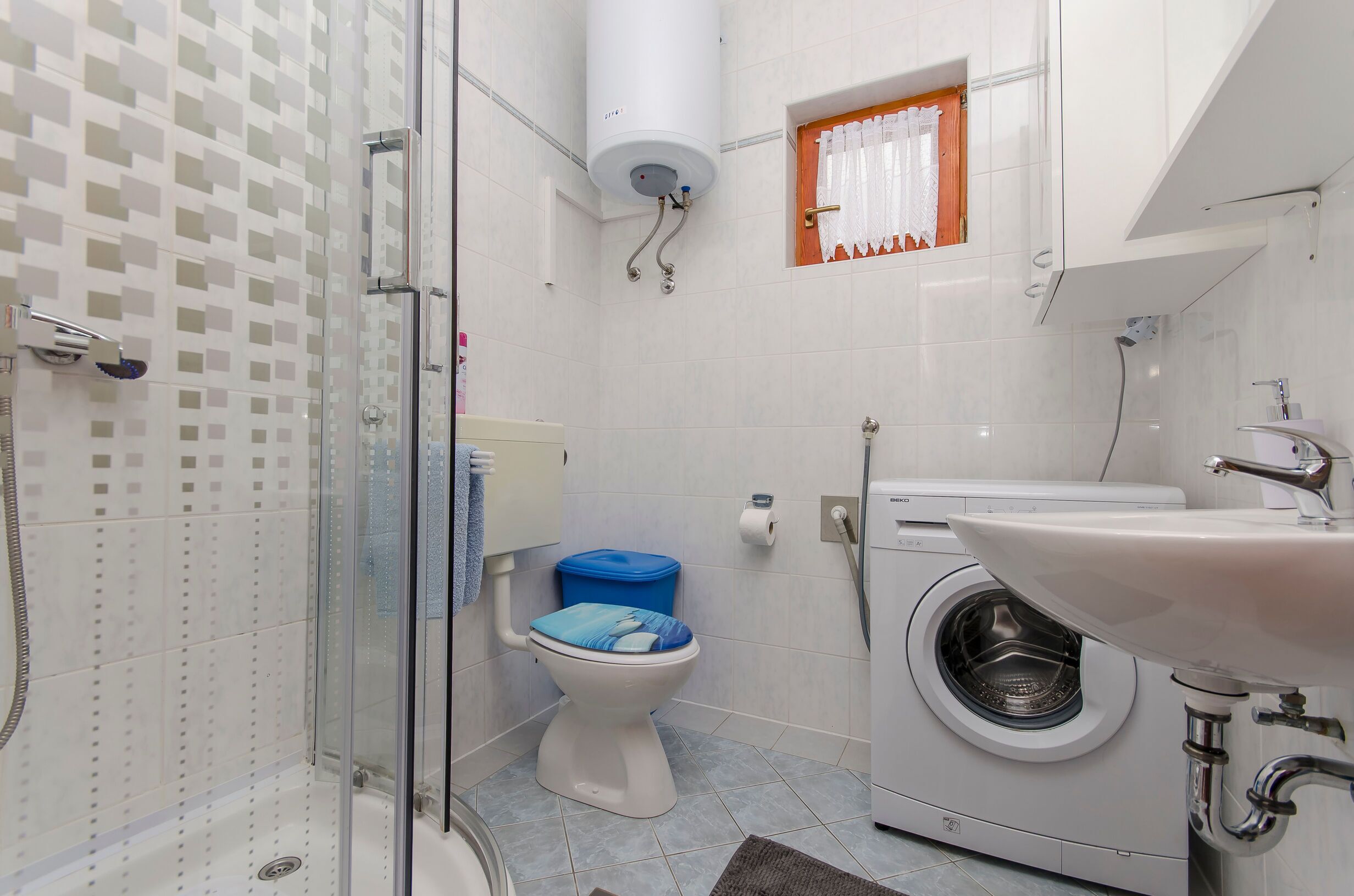 A bathroom with a glass shower enclosure, a toilet with a blue lid, a washing machine, and a sink.