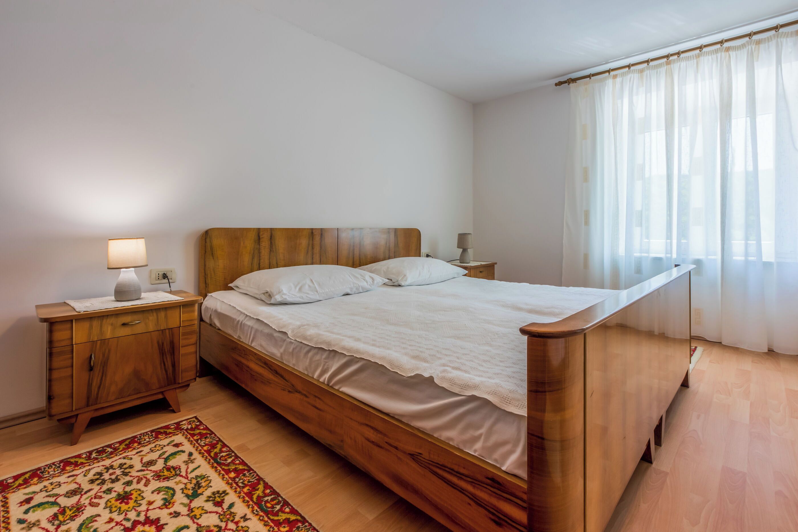 A bedroom with a wooden bed, a nightstand, a lamp, and a window with curtains.
