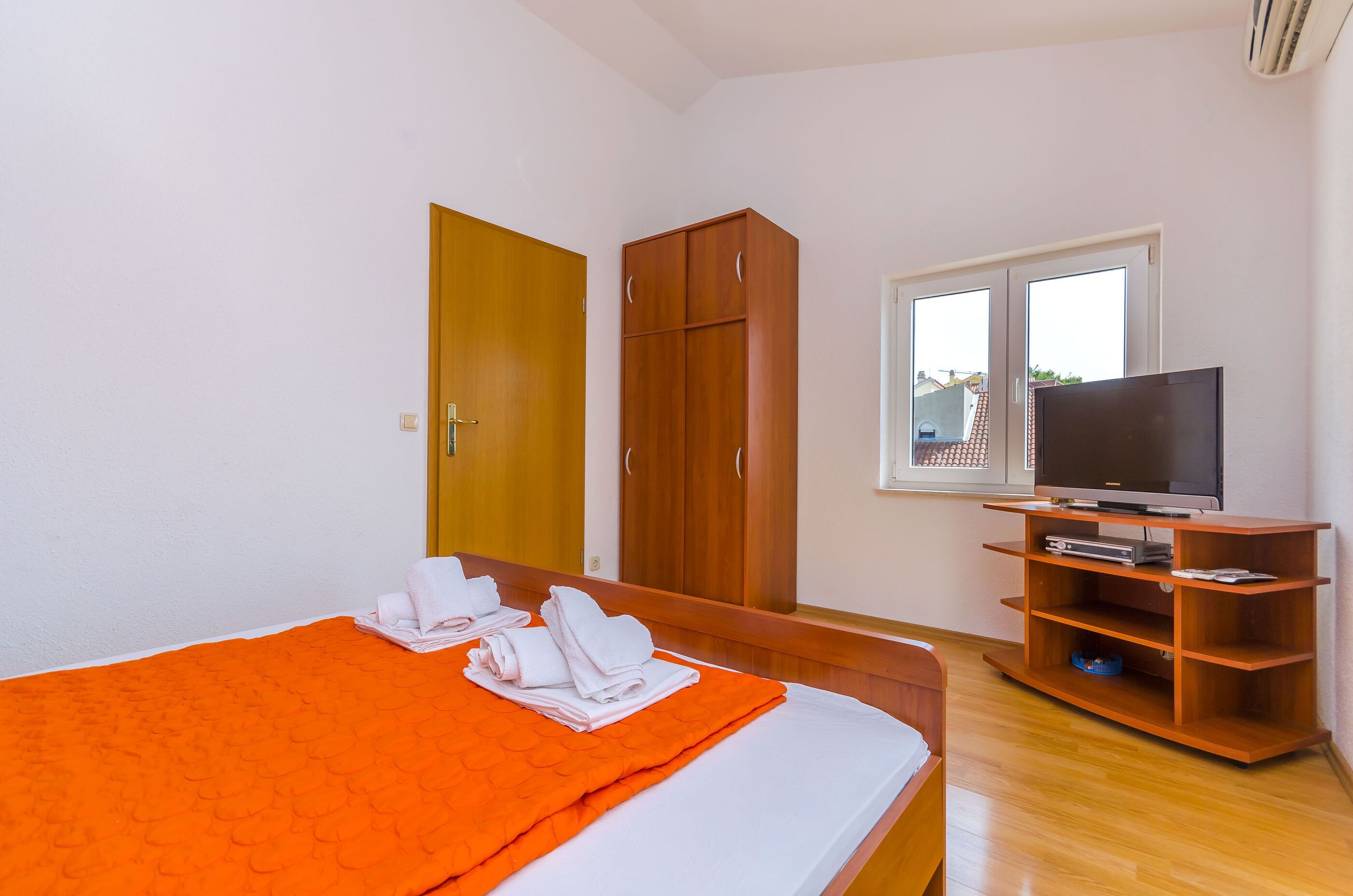 A hotel room with a bed, a wooden wardrobe, a television on a wooden stand, and a window with a view of buildings.