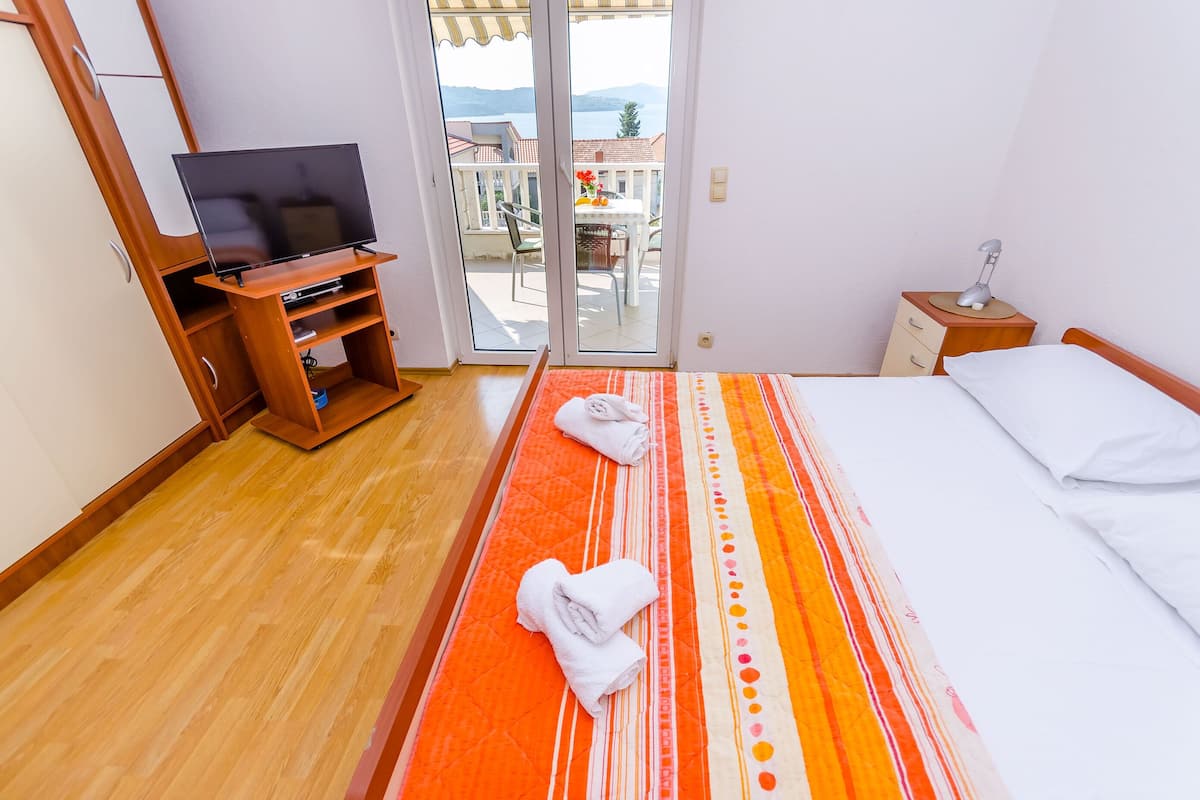 A hotel room with a bed, a TV on a wooden stand, a small table with a lamp, and a view of the outside balcony.