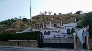 Una casa de dos pisos con balcón, entrada protegida por un portón y un paisaje montañoso de fondo.