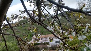 Un edificio con fachada blanca y ventanas arqueadas, rodeado de un paisaje montañoso y árboles en flor.