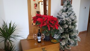 Un aparador de madera con una botella de vino, dos copas y una planta en maceta. Un árbol de Navidad con adornos blancos está al lado del aparador.