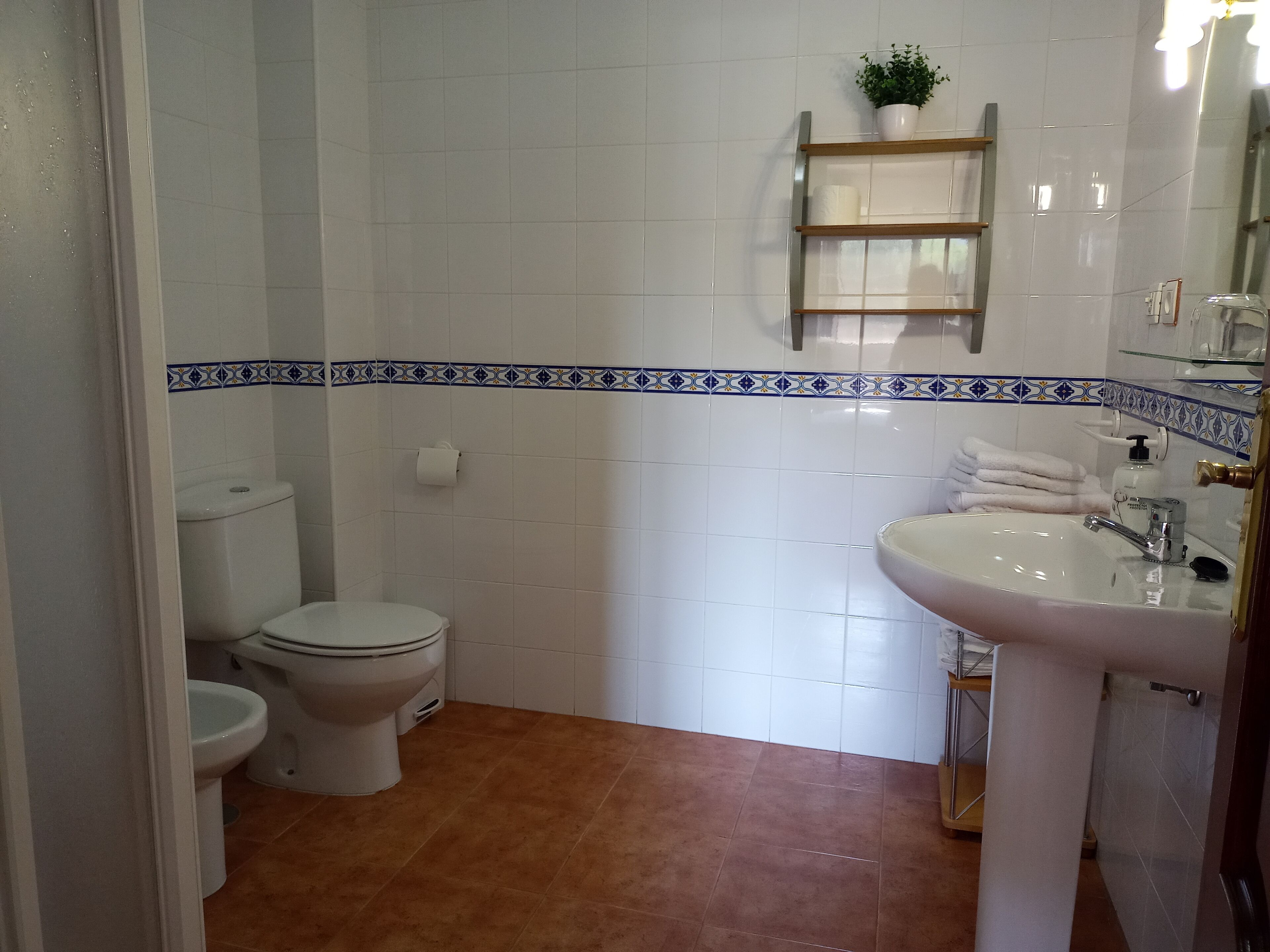 A bathroom with white tiled walls, a brown floor, a white sink with a gold faucet, a white toilet, and a shelf with a plant.