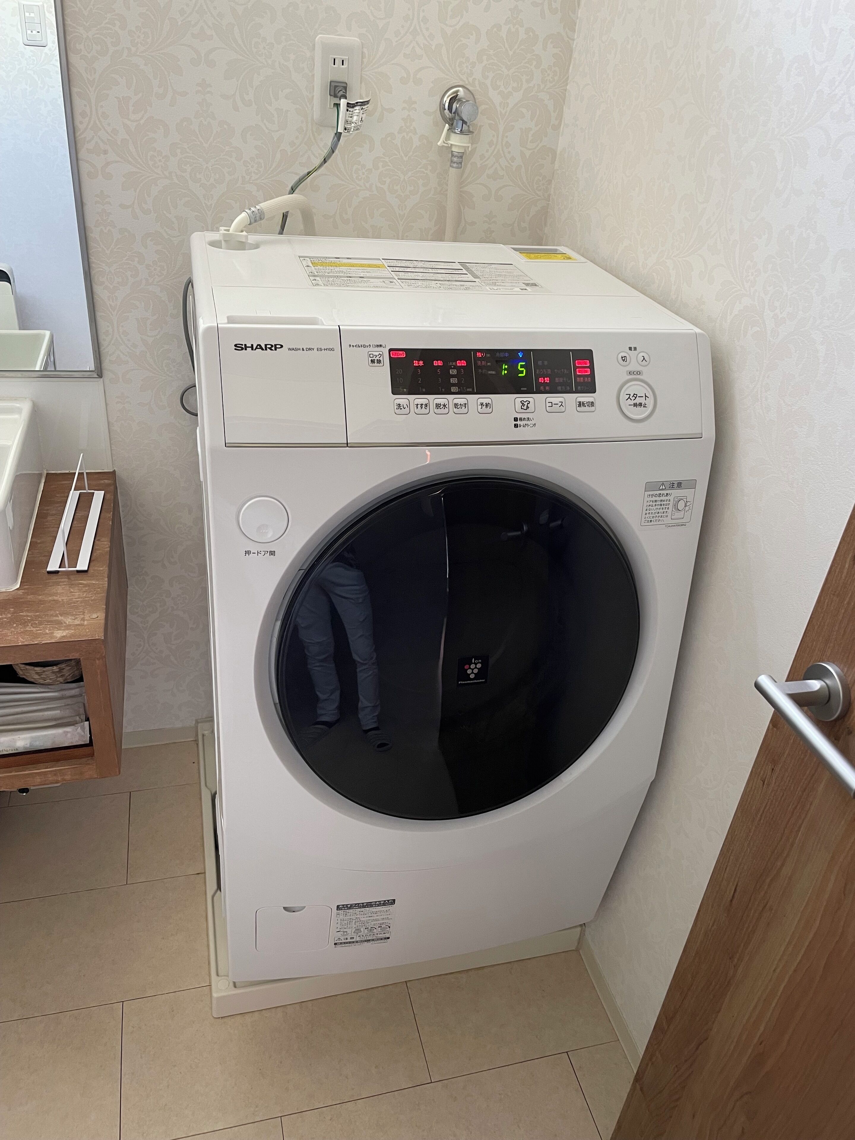 Eine kompakte, weiße Waschmaschine mit Frontlader und digitaler Anzeige, platziert in einer Ecke eines Zimmers mit strukturierter Tapete und einem Holzschrank.